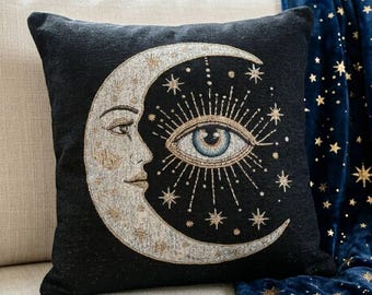 Pode incluir: Uma almofada quadrada preta com um design tecido de uma lua crescente com perfil de rosto humano. A lua é representada em tons de prata e ouro, com um olho azul central rodeado por linhas radiantes e pequenas estrelas douradas.