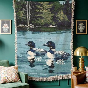 May include: A woven throw blanket featuring a detailed nature scene of two loons swimming on a calm blue lake with a dark green forest shoreline in the background. The rectangular textile has a cream-coloured fringe along the vertical edges.