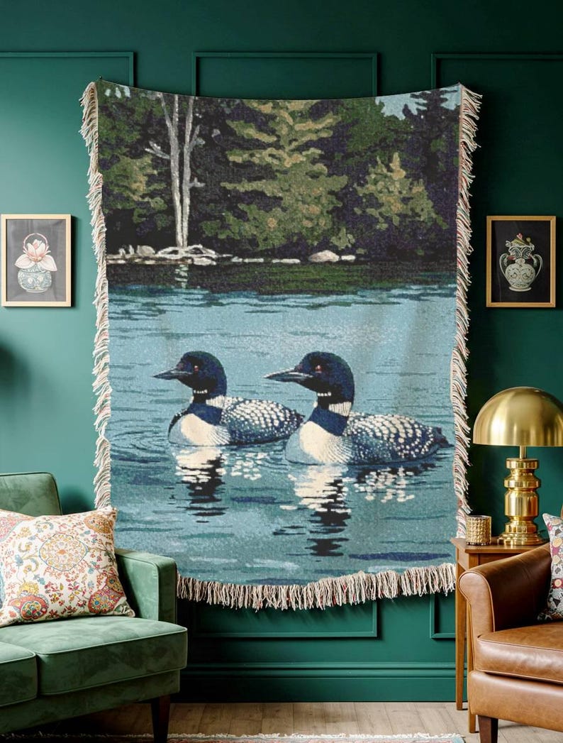 May include: A woven throw blanket featuring a detailed nature scene of two loons swimming on a calm blue lake with a dark green forest shoreline in the background. The rectangular textile has a cream-coloured fringe along the vertical edges.