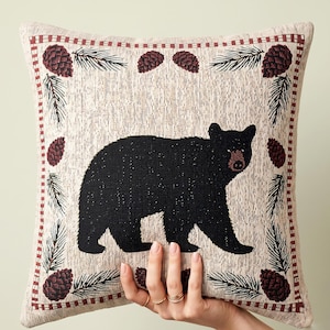 May include: A square throw pillow featuring a woven tapestry design of a black bear centered on a light beige background. The border is decorated with a repeating pattern of dark red pinecones and green pine needles framed by a thin red line.