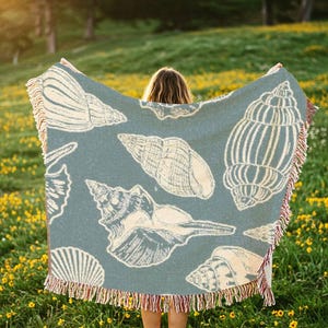 Puede incluir: Una persona sosteniendo una manta tejida azul y blanca con un patr&oacute;n de conchas marinas. La manta tiene flecos en los bordes. La persona est&aacute; de pie en un campo de flores amarillas.