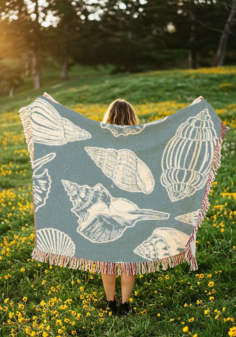 Puede incluir: Una persona sosteniendo una manta tejida azul y blanca con un patr&oacute;n de conchas marinas. La manta tiene flecos en los bordes. La persona est&aacute; de pie en un campo de flores amarillas.