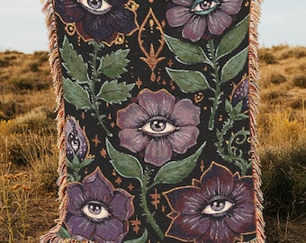 Könnte beinhalten: Eine gewebte Wandteppich-Decke mit dunklem Hintergrund und einem sich wiederholenden Muster aus lila Blüten mit Augen. Die Blüten sind von goldenen Sternen und grünen Blättern umgeben.
