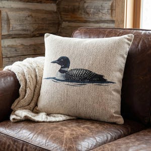 May include: A square throw pillow featuring a textured beige fabric with a detailed illustration of a black and white loon swimming on water. The bird is depicted in profile, showcasing its characteristic spotted plumage and dark head against the neutral background.