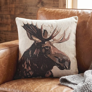 May include: A square throw pillow featuring a woven tapestry design of a moose head in profile. The moose is rendered in shades of dark brown and black against a light beige background, showcasing detailed antlers and a textured fabric finish.