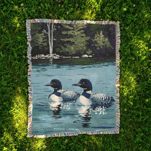 May include: A rectangular woven throw blanket featuring a detailed scene of two loons swimming on a calm lake. The background displays a dark green forest shoreline with trees. The blanket has a multi-coloured fringed edge on all four sides.