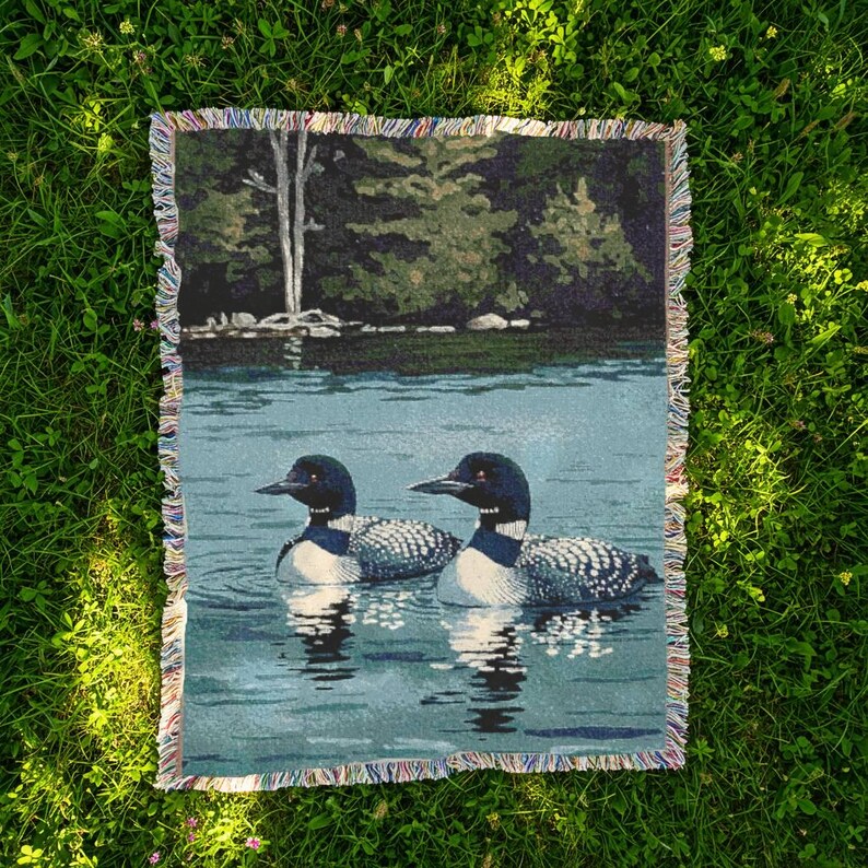 May include: A rectangular woven throw blanket featuring a detailed scene of two loons swimming on a calm lake. The background displays a dark green forest shoreline with trees. The blanket has a multi-coloured fringed edge on all four sides.