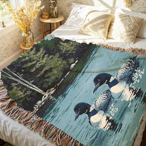 May include: A woven throw blanket featuring a detailed scene of two loons swimming on a calm blue lake next to a dark green shoreline with trees. The blanket has a textured appearance and is finished with a multi-coloured fringed edge on all sides.