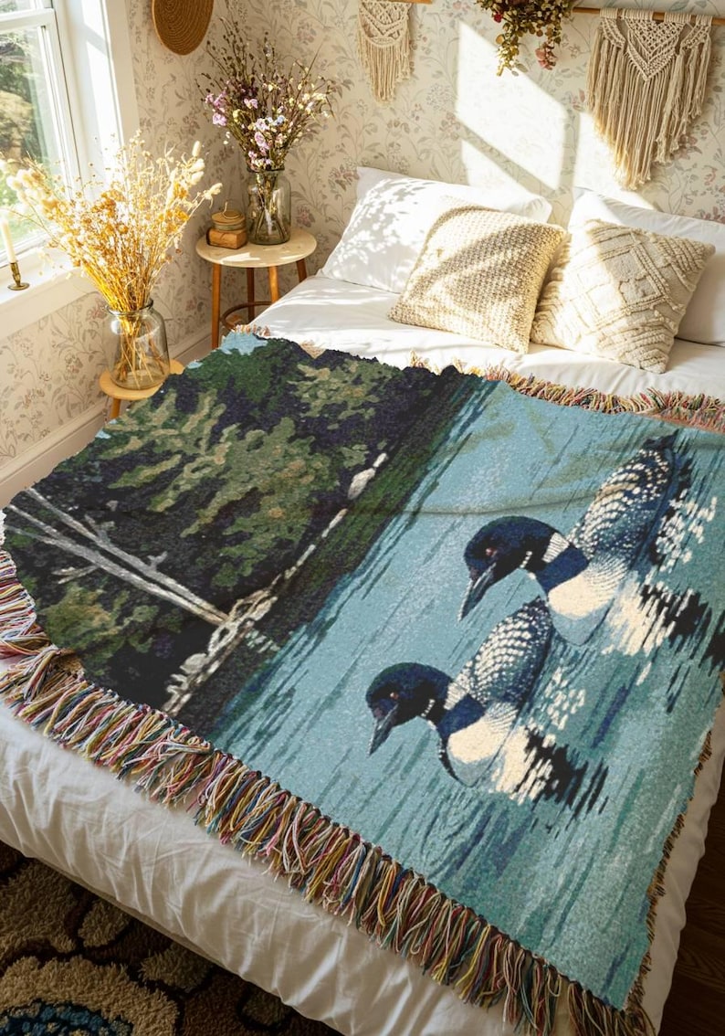 May include: A woven throw blanket featuring a detailed scene of two loons swimming on a calm blue lake next to a dark green shoreline with trees. The blanket has a textured appearance and is finished with a multi-coloured fringed edge on all sides.