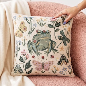 May include: A square throw pillow with a cream-colored fabric base featuring a woven tapestry design of a central green frog surrounded by various insects including butterflies, a dragonfly, moths, and ladybugs, interspersed with colorful wildflowers.