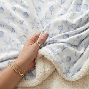 May include: A white blanket patterned with blue seashells and starfish. The blanket has a soft, plush texture with a cream-coloured sherpa lining. The design features various seashell and starfish illustrations.