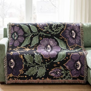 Könnte beinhalten: Eine gewebte Wohndecke mit schwarzem Hintergrund und einem sich wiederholenden Muster aus violetten Blumen, die jeweils ein zentrales Auge enthalten. Das Design umfasst grüne Blätter, goldfarbene Sternakzente und dekorative Fransen an den Rändern.