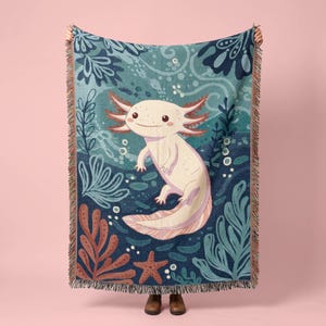 May include: A woven throw blanket featuring a central illustration of a light pink axolotl surrounded by dark blue water, teal aquatic plants, and a brown starfish. The blanket has a fringed edge and a detailed, textured tapestry-style weave.