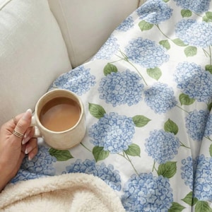 May include: A cream-colored mug filled with a light brown beverage rests on a white and blue floral blanket. The blanket features a pattern of blue hydrangea flowers and green leaves. A hand with rings holds the mug, suggesting a cozy, relaxing scene.