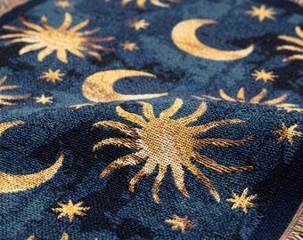 Puede incluir: Una manta tejida con un fondo azul profundo y un patrón repetitivo de soles, lunas crecientes y estrellas de color amarillo dorado. La tela presenta un tejido texturizado tipo tapiz con flecos visibles en los bordes superior e inferior.