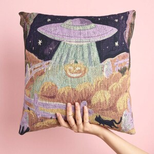 Puede incluir: Un cojín cuadrado con un diseño de tapiz tejido que muestra un OVNI morado sobre un campo de calabazas por la noche. La escena incluye una calabaza iluminada, una valla de madera, pequeñas figuras de fantasmas y un gato negro.