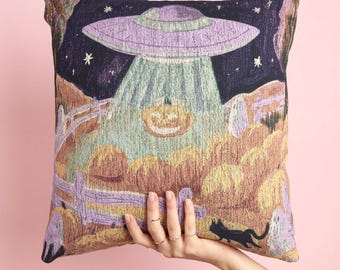 Può includere: Un cuscino quadrato con un design a arazzo intrecciato che raffigura un UFO viola che sorvola un campo di zucche di notte. La scena include una zucca illuminata, una staccionata in legno, piccoli fantasmi e un gatto nero su sfondo scuro.