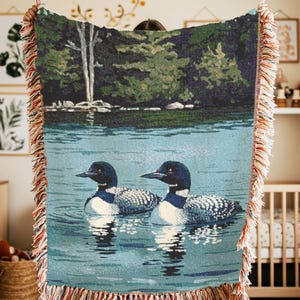 May include: A woven throw blanket featuring a detailed scene of two loons swimming on a calm blue lake with a dark green forest and a white tree in the background. The blanket has a decorative fringe edge with multi-coloured threads throughout.
