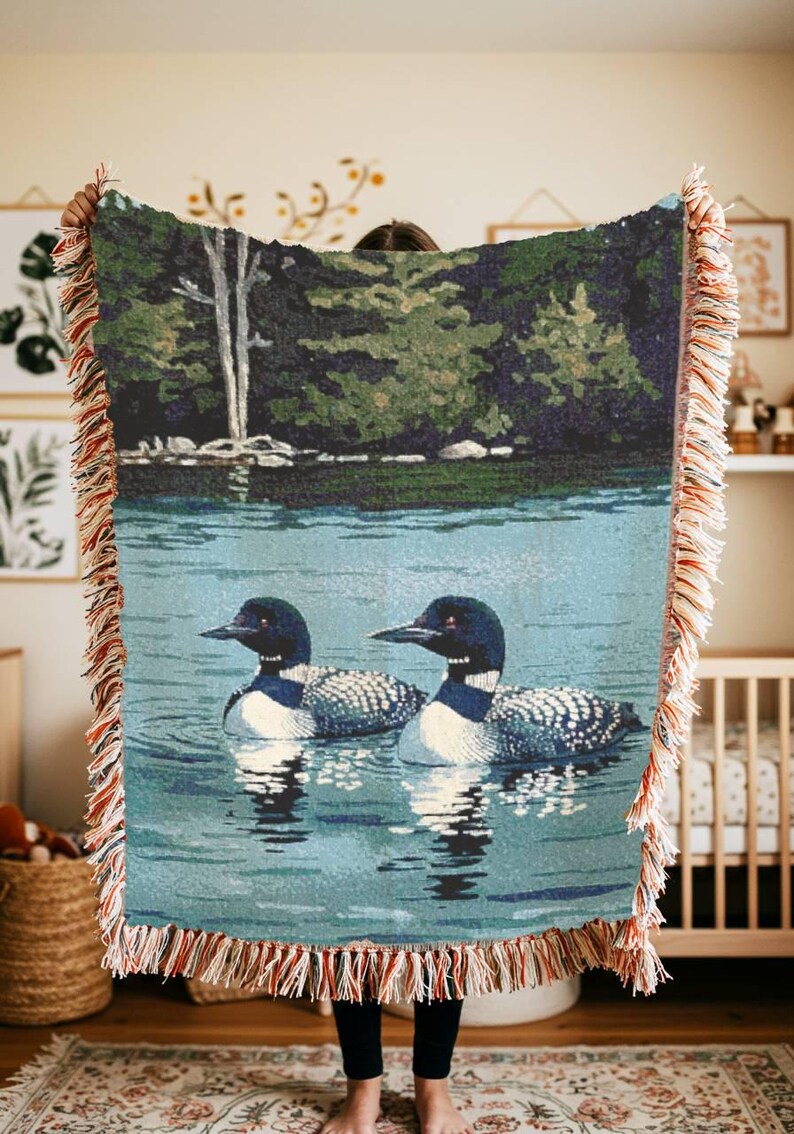 May include: A woven throw blanket featuring a detailed scene of two loons swimming on a calm blue lake with a dark green forest and a white tree in the background. The blanket has a decorative fringe edge with multi-coloured threads throughout.
