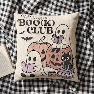 May include: A square beige throw pillow featuring the text Midnight BOO(K) CLUB in black. The design includes two white ghosts reading purple books, two orange pumpkins, a black cat sitting on a stack of books, a small brown mug, and two black bats.