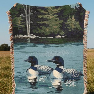 May include: A woven throw blanket featuring a detailed scene of two loons swimming on a calm blue lake. The background depicts a dense green forest with a tall white tree. The blanket has a decorative fringed edge with multi-coloured threads.