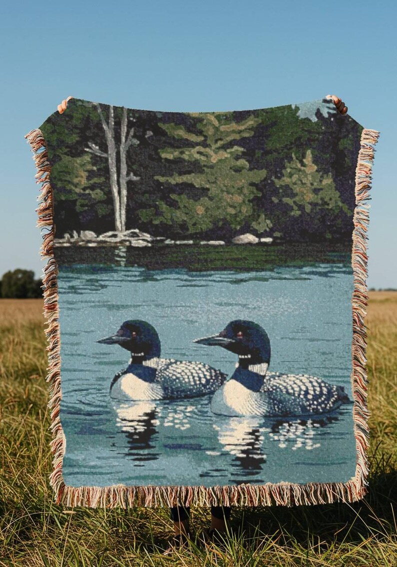 May include: A woven throw blanket featuring a detailed scene of two loons swimming on a calm blue lake. The background depicts a dense green forest with a tall white tree. The blanket has a decorative fringed edge with multi-coloured threads.