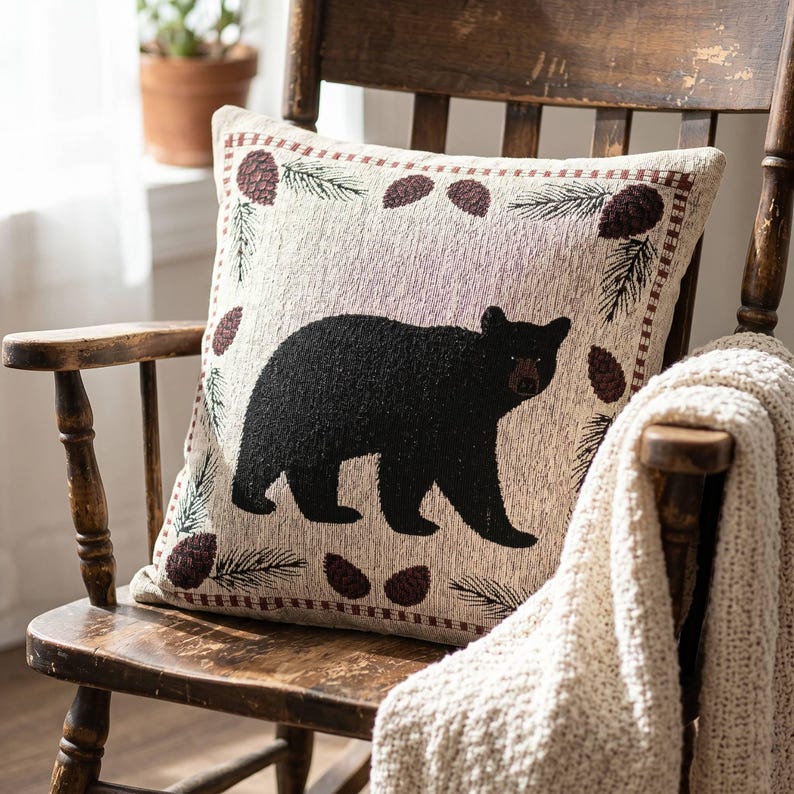 May include: A square throw pillow featuring a woven design of a solid black bear centered on a light beige background. The border is decorated with a repeating pattern of dark brown pinecones and green pine needles in a rectangular frame.