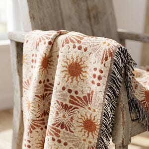 Puede incluir: Una manta tejida con un patrón repetitivo de soles y formas geométricas en tonos naranja óxido y crema. La manta tiene una superficie texturizada y está terminada con flecos decorativos a lo largo de los bordes, mostrando un diseño detallado.