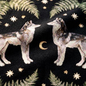 May include: A black woven textile featuring two wolves facing each other, centred by a gold crescent moon. The design includes green fern leaves and scattered gold stars across the dark fabric, creating a detailed, repeating pattern.
