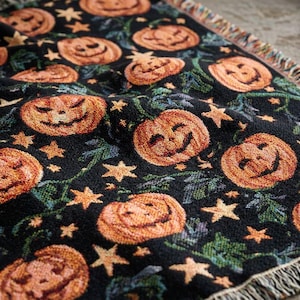 May include: A black woven throw blanket featuring a repeating pattern of orange jack-o'-lanterns with carved faces, interspersed with small yellow stars and green vine details. The blanket has a textured surface and a fringed edge along the side.