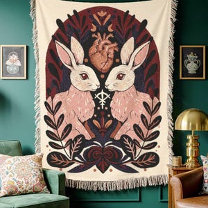 May include: A cream throw blanket with a symmetrical design of two pink rabbits facing each other, with a heart and floral elements. The artwork is framed by a dark burgundy arch and fringed edges.