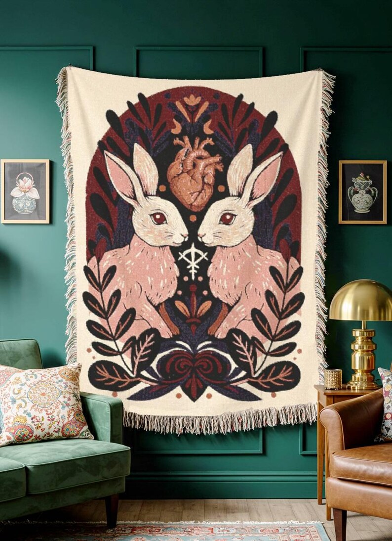 May include: A cream throw blanket with a symmetrical design of two pink rabbits facing each other, with a heart and floral elements. The artwork is framed by a dark burgundy arch and fringed edges.