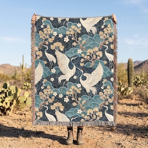 May include: A rectangular woven throw blanket featuring a repeating pattern of white cranes, golden-brown pine trees, and teal wave motifs against a dark blue background. The blanket has a fringed edge on the top and bottom sides.