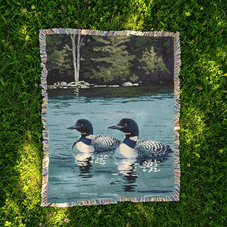 May include: A rectangular woven throw blanket featuring a detailed scene of two loons swimming on a calm lake. The background displays a dark green forest shoreline with trees. The blanket has a multi-coloured fringed edge on all four sides.