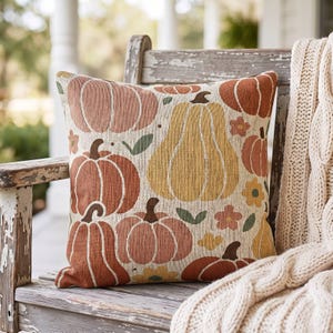 Pode incluir: Uma almofada quadrada com um padrão tecido de abóboras em tons de laranja, amarelo e rosa suave, intercalado com pequenas flores e folhas verdes sobre um fundo creme. O tecido apresenta uma textura visível e entrelaçada.