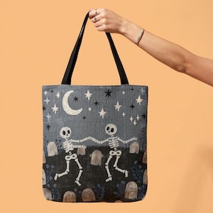 May include: A rectangular tote bag with black handles featuring a woven design of two white skeletons dancing in a graveyard at night. The scene includes a crescent moon, various stars, and several tombstones set against a dark gray background.