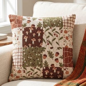 Könnte beinhalten: Ein quadratisches Zierkissen mit Patchwork-Design und verschiedenen herbstlichen Mustern. Der Stoff zeigt Illustrationen von braunen Pilzen, orangefarbenen Kürbissen, grünen Eichenblättern und Eicheln auf cremefarbenen, dunkelbraunen und olivgrünen Hintergründen.