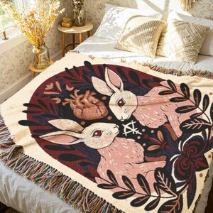May include: A woven throw blanket with a design of two rabbits with red eyes, surrounded by floral and botanical elements. The colour palette includes cream, burgundy, and black. The blanket has a fringed edge.