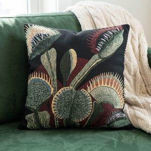 May include: A square throw pillow featuring a woven tapestry design of multiple Venus flytrap plants. The botanical illustration displays green and reddish-brown leaves with spiky edges against a solid black background, creating a detailed texture.