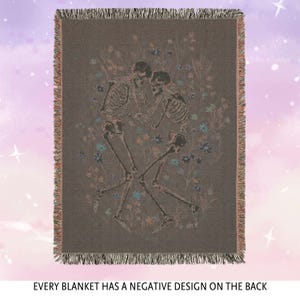 May include: A woven tapestry throw blanket with a dark brown background featuring a design of three skeletons dancing in a field of flowers. The skeletons are outlined in a light pink color and the flowers are in shades of blue, pink, and yellow.
