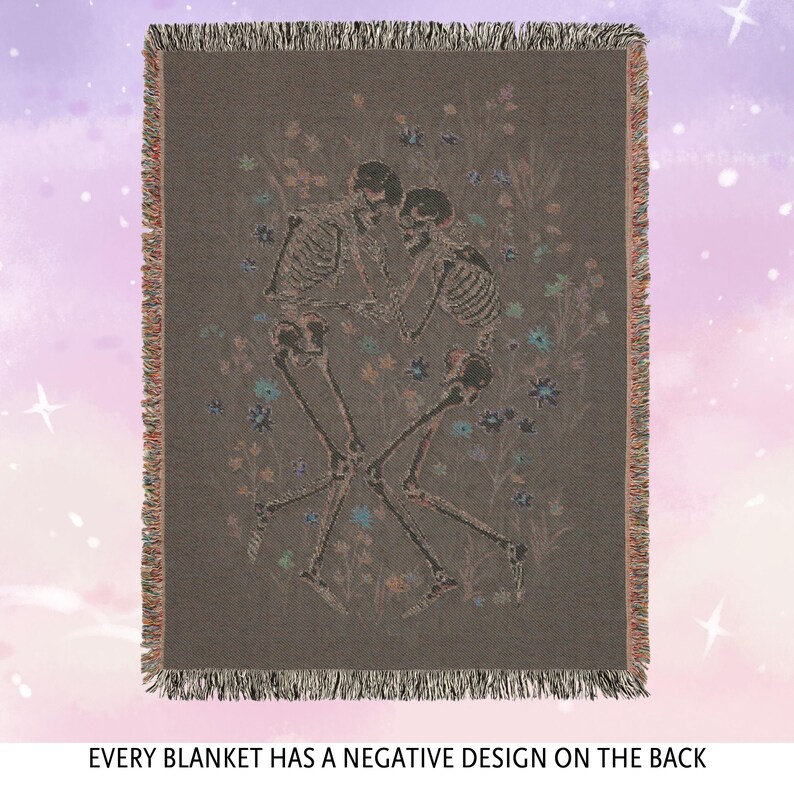 May include: A woven tapestry throw blanket with a dark brown background featuring a design of three skeletons dancing in a field of flowers. The skeletons are outlined in a light pink color and the flowers are in shades of blue, pink, and yellow.