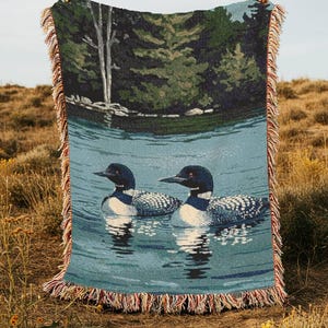 May include: A woven throw blanket featuring a detailed scene of two loons swimming on a calm blue lake. The background depicts a dense forest with green trees and a white birch trunk. The blanket has a decorative fringe edge with multi-coloured threads.