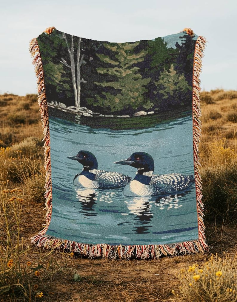 May include: A woven throw blanket featuring a detailed scene of two loons swimming on a calm blue lake. The background depicts a dense forest with green trees and a white birch trunk. The blanket has a decorative fringe edge with multi-coloured threads.