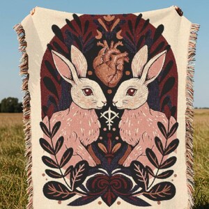May include: A cream throw blanket with a design of two pink rabbits, a brown heart, and botanical motifs. The blanket has a fringed edge. The design is set against a dark background.