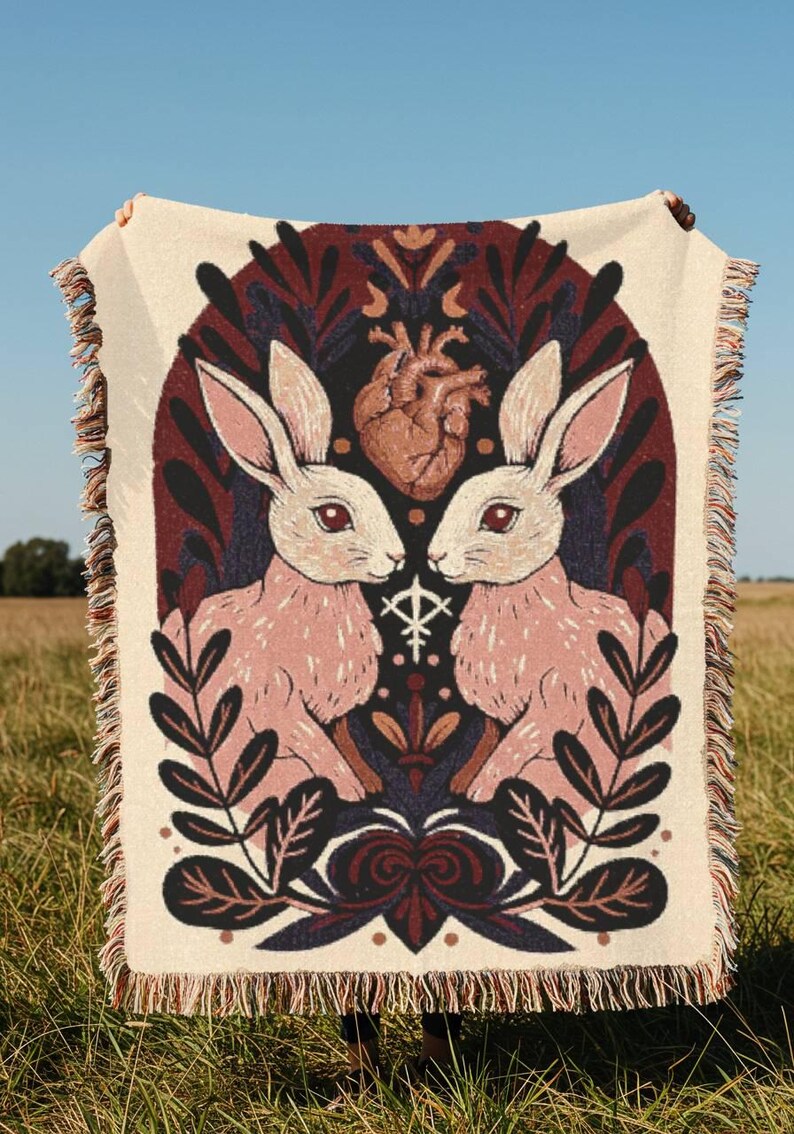 May include: A cream throw blanket with a design of two pink rabbits, a brown heart, and botanical motifs. The blanket has a fringed edge. The design is set against a dark background.