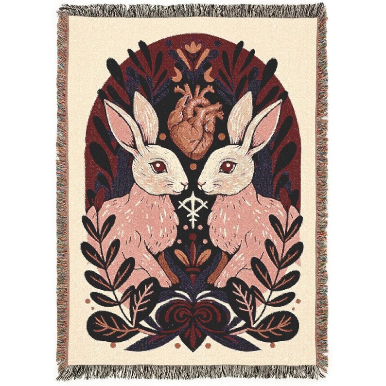 May include: Cream-coloured woven throw blanket featuring a design with two pink rabbits facing each other, a brown heart, and botanical elements. The design is framed by a dark red arch and includes a fringe border.