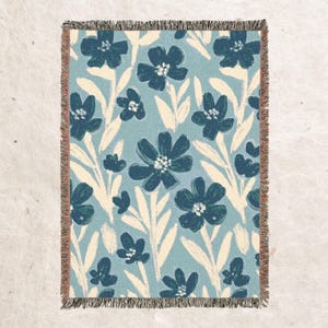 May include: A woven throw blanket with a blue and white floral pattern. The blanket features a repeating pattern of stylised flowers with white stems and leaves on a light blue background.