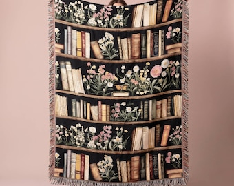 May include: A black woven throw blanket featuring a repeating pattern of wooden bookshelves filled with various books and colorful floral arrangements. The blanket has a fringed edge on the top and bottom and a dark, detailed design throughout.