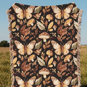 Könnte beinhalten: Eine schwarze Decke mit einem sich wiederholenden Muster aus Schmetterlingen, Pilzen und Blättern in Braun-, Beige- und Cremetönen. Die Decke hat einen Fransenrand und wird im Freien vor einem Feld und blauem Himmel präsentiert.