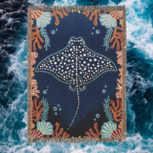 Puede incluir: Una manta de tejido de tapicería con una raya azul manchada nadando en un océano azul oscuro con corales y conchas marinas. La manta tiene un borde con flecos.